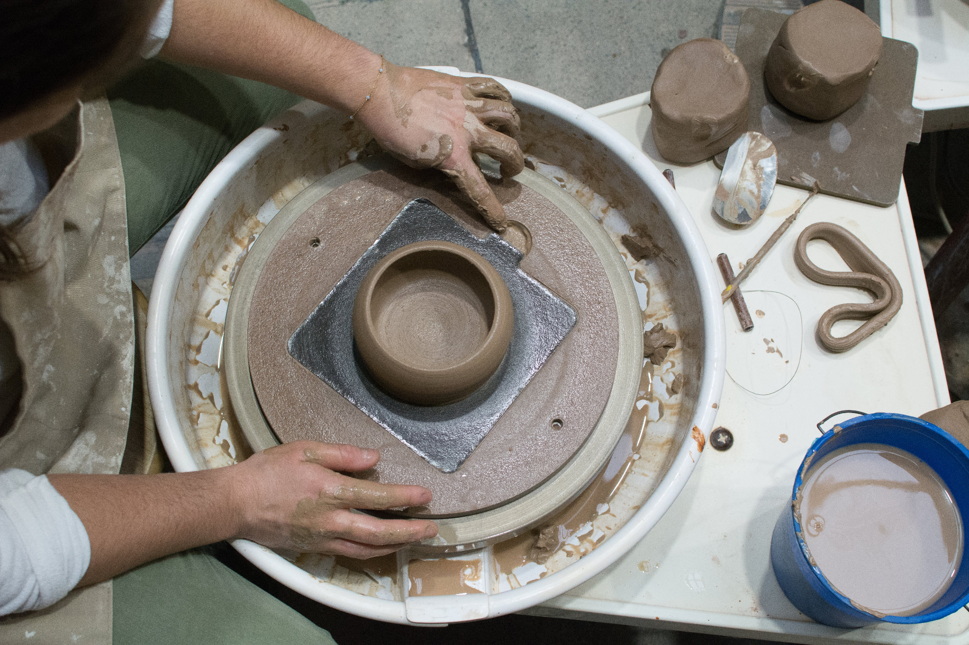 Top-down view of a wheel-thrown cup in progress beside trimming tools and slip bucket